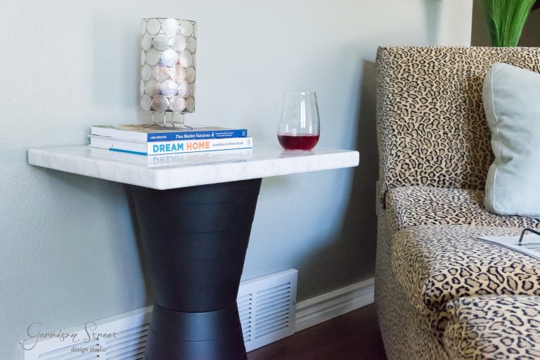 A small side table with a marble top holds a stack of books and a half-full glass of red wine. The table is next to a leopard-print sofa, with a candle holder adding to the cozy atmosphere.