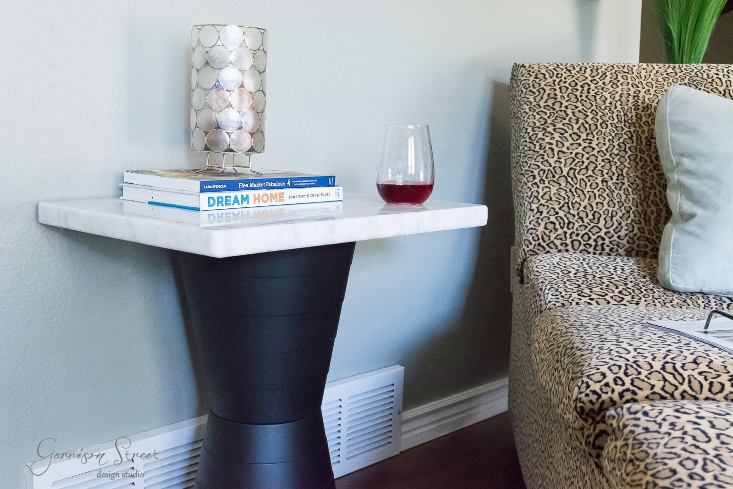 A small side table with a marble top holds a stack of books and a half-full glass of red wine. The table is next to a leopard-print sofa, with a candle holder adding to the cozy atmosphere.