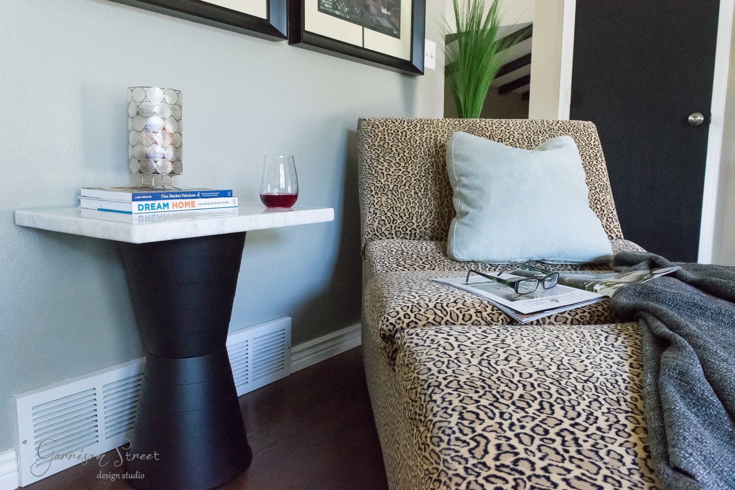 A cozy room corner featuring a leopard print chaise lounge with a light blue pillow. A small round table holds a red wine glass and books. A fluffy gray blanket is draped over the chaise. A framed picture and green plant add decor in the background.