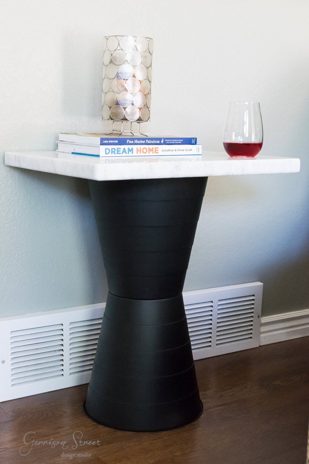 A small side table with a black hourglass base and white marble top. It holds three books, a decorative candle, and a glass of red wine. The table is on a wooden floor next to a wall vent.