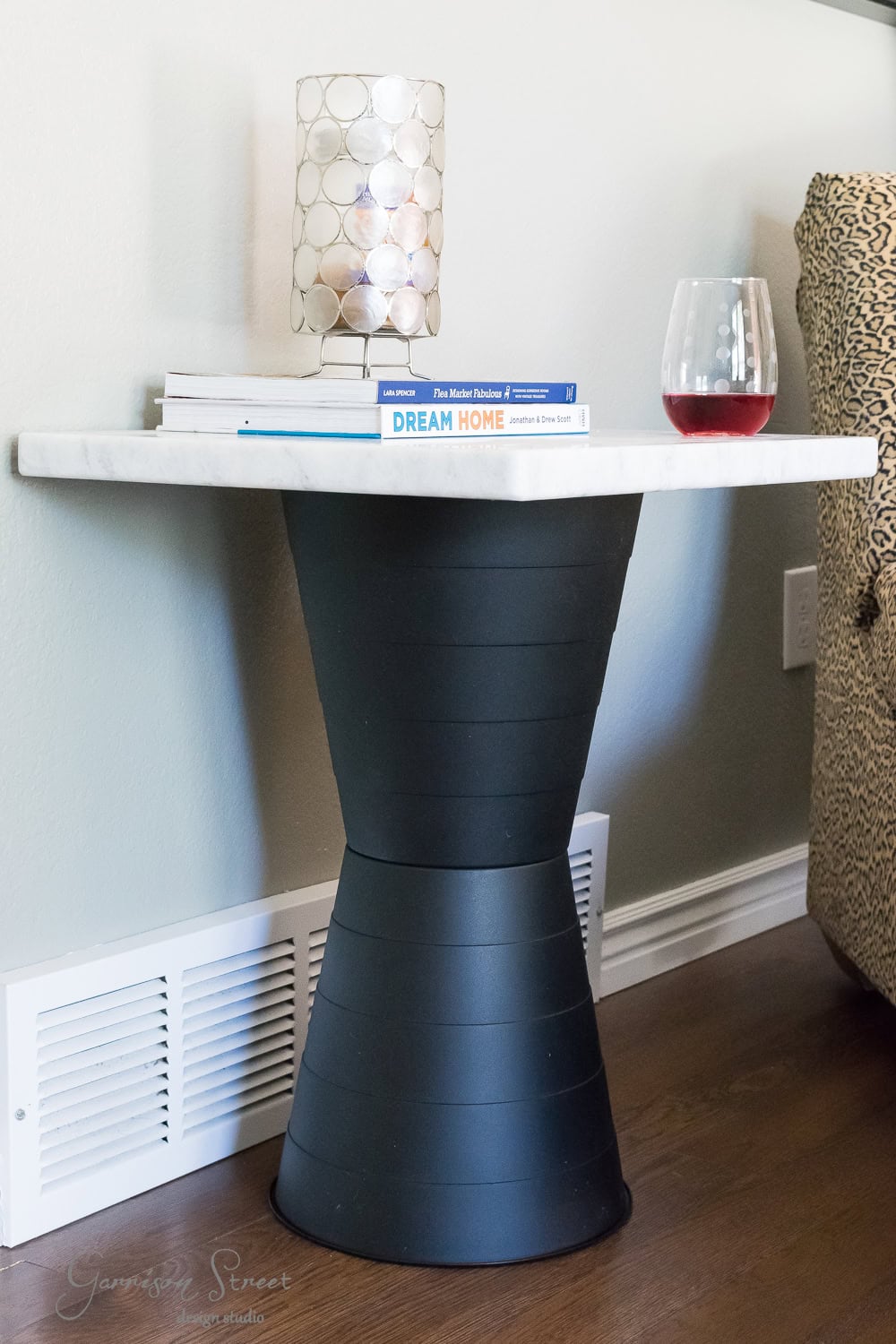 A modern side table with a white marble top and a black hourglass-shaped base. On the table are a stack of books, a decorative glass candle holder, and a glass of red wine. The table is placed on a wooden floor next to a patterned chair.