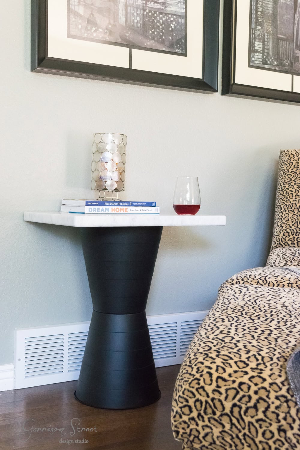 A modern living room scene featuring a stylish side table with a black base and white marble top. On the table are a decorative candle holder, an open book, and a wine glass. Nearby is a leopard-print chair and framed artwork on the wall.