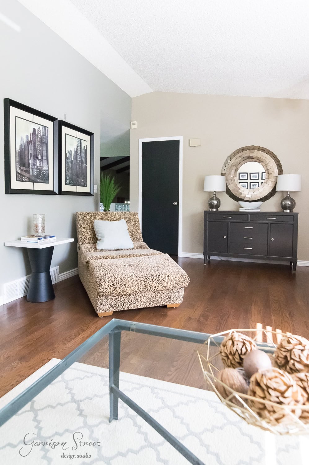 A modern living room features a leopard print lounge chair, a black side table, and framed cityscape art on the wall. A round mirror and two lamps sit atop a dark sideboard. The room has wooden floors and neutral-toned walls.