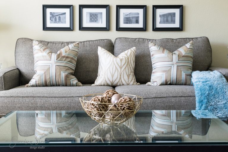 A cozy living room with a gray sofa adorned with patterned pillows under a series of framed black-and-white photos. A glass coffee table in the foreground holds a decorative metal bowl filled with rattan balls.