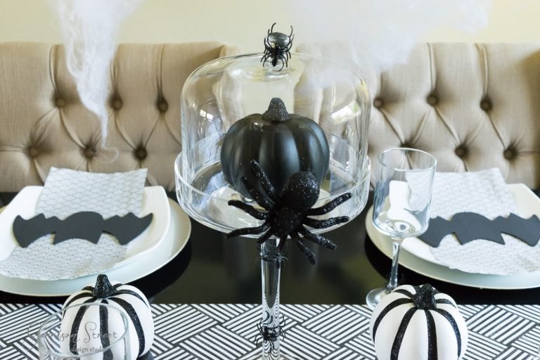 A Halloween-themed dining table setup features a black pumpkin under a glass cloche embellished with spooky spider decorations. Plates with bat-shaped napkins and white pumpkins with black stripes complete the monochrome decor.