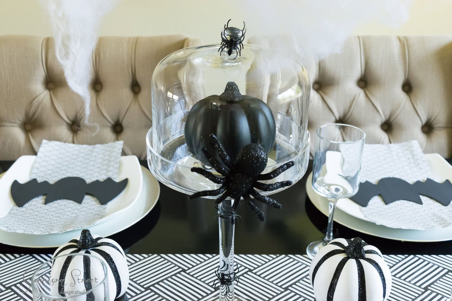 A Halloween-themed dining table setup features a black pumpkin under a glass cloche embellished with spooky spider decorations. Plates with bat-shaped napkins and white pumpkins with black stripes complete the monochrome decor.