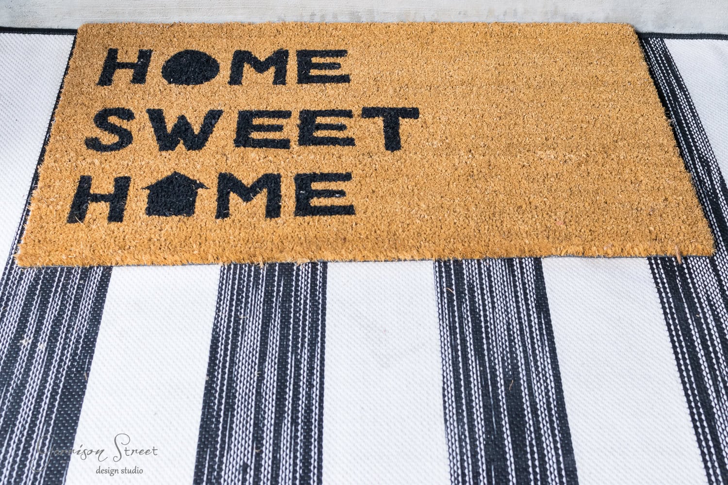 A doormat with the words "Home Sweet Home" written in black on a brown background. The mat is placed on a striped black and white rug.