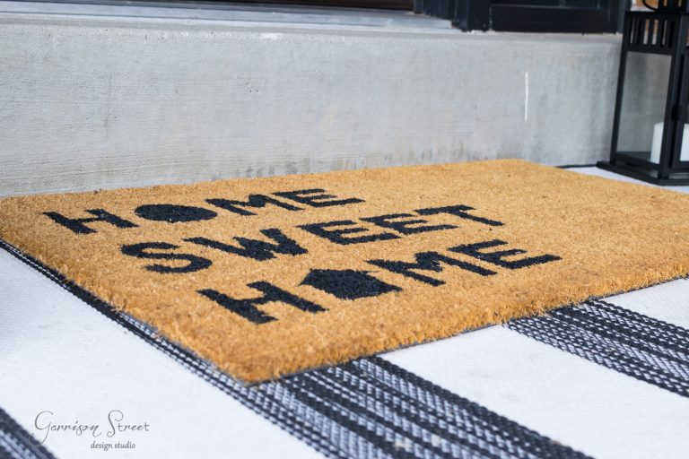 A brown doormat with the black text "HOME SWEET HOME" placed on a black and white striped rug near a doorway. A black lantern is visible to the right.