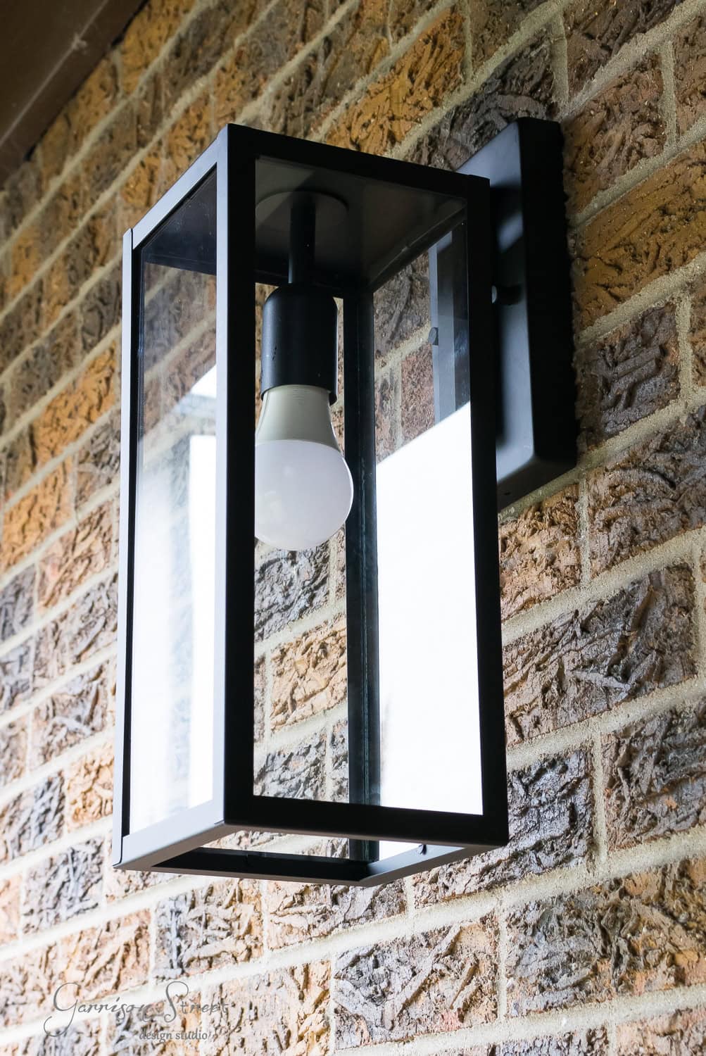 A modern black metal and glass outdoor wall lantern mounted on a brick wall. The light fixture contains a visible light bulb. The bricks have a textured, multicolored pattern.