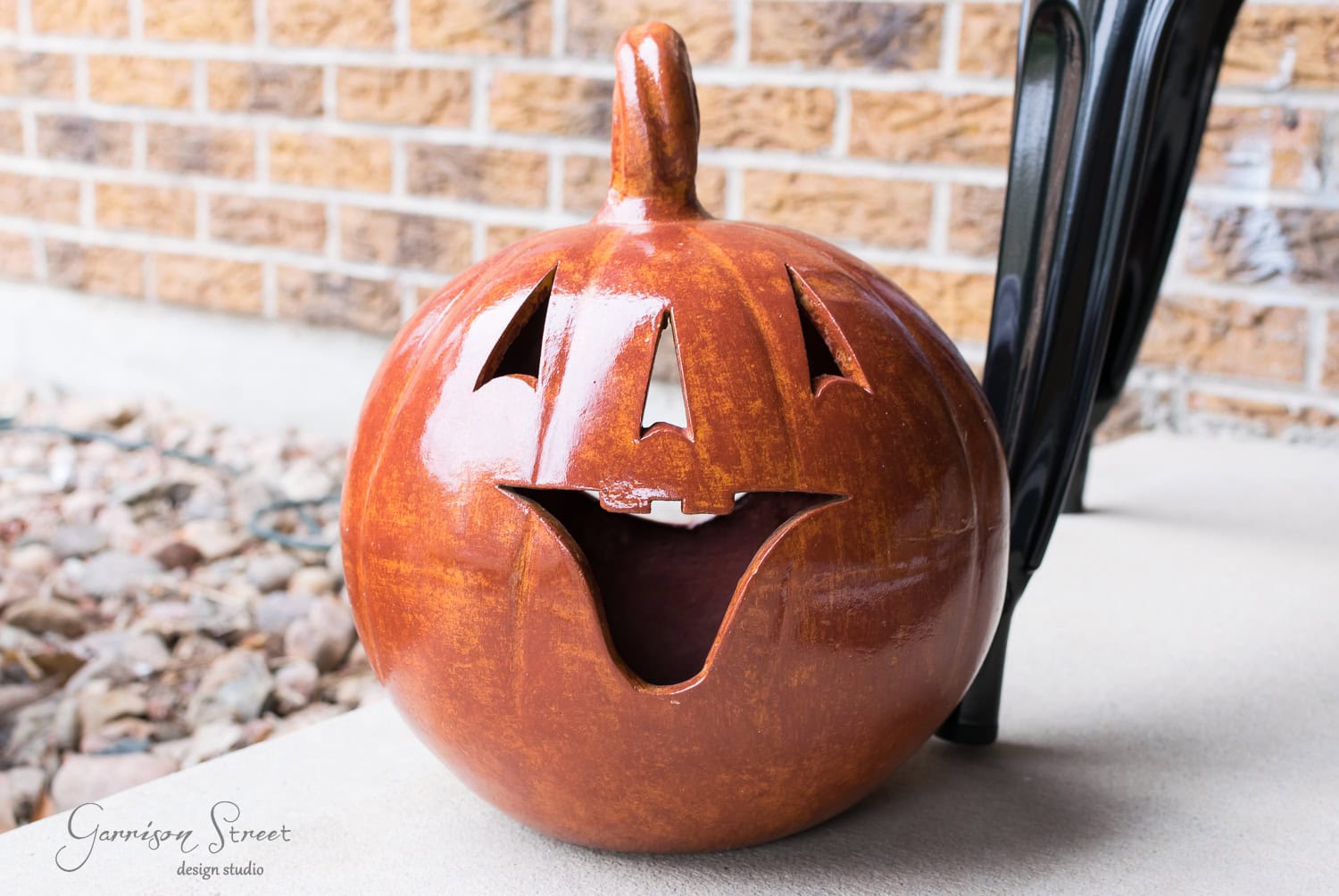 A ceramic pumpkin with a carved, smiling face sits on a porch. The pumpkin is a deep orange color and has a glossy surface. Behind it, a brick wall and a portion of black furniture are visible.