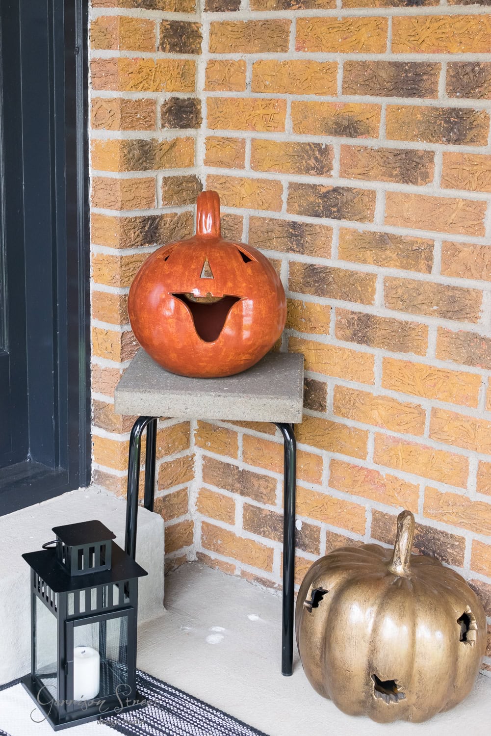 A carved pumpkin with a happy face sits on a small table against a brick wall. Below, a gold pumpkin rests on the ground. A black lantern with a candle is placed nearby on a doormat.