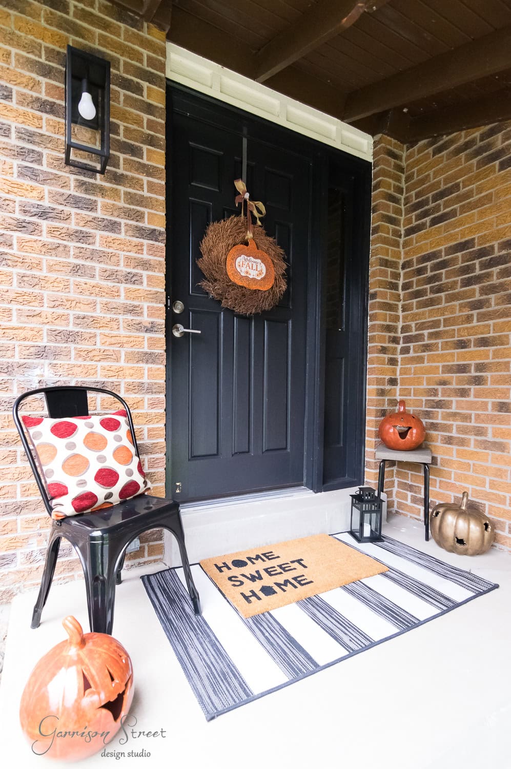 A cozy front porch decorated for fall with a black door and a wreath. There's a doormat with "Home Sweet Home," metal chair with a patterned cushion, lantern, and several pumpkins in orange and silver hues on the step.