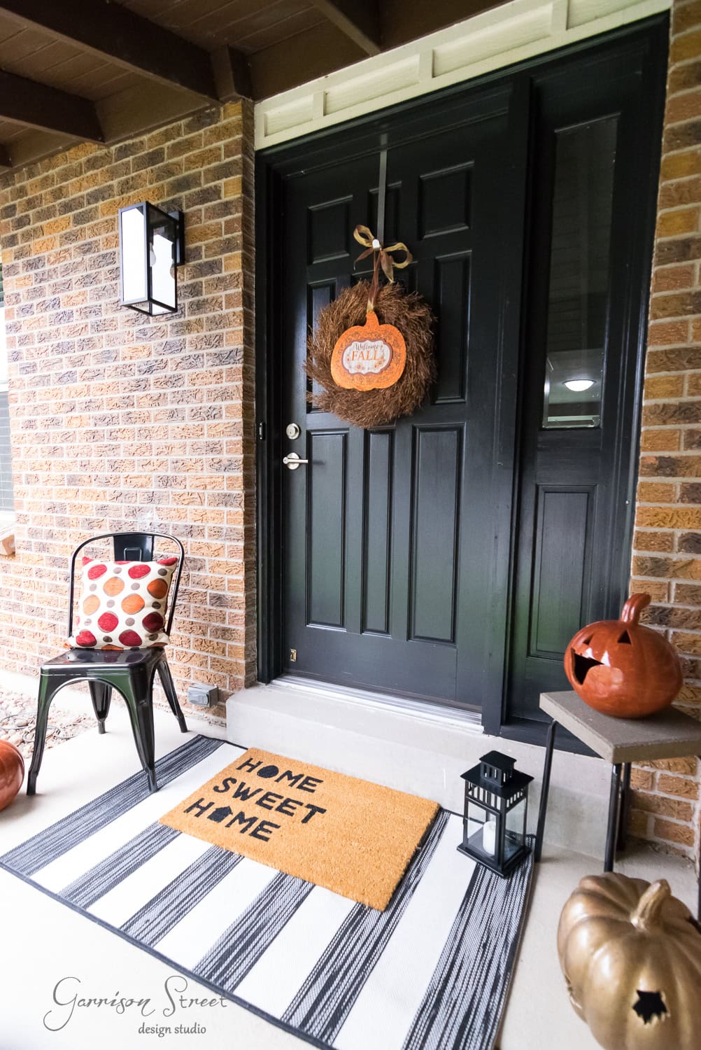 Front porch decorated for fall with a black door and brick walls. A "Home Sweet Home" doormat, lantern, pumpkins, and a chair with a polka dot cushion adorn the area. A festive wreath hangs on the door.
