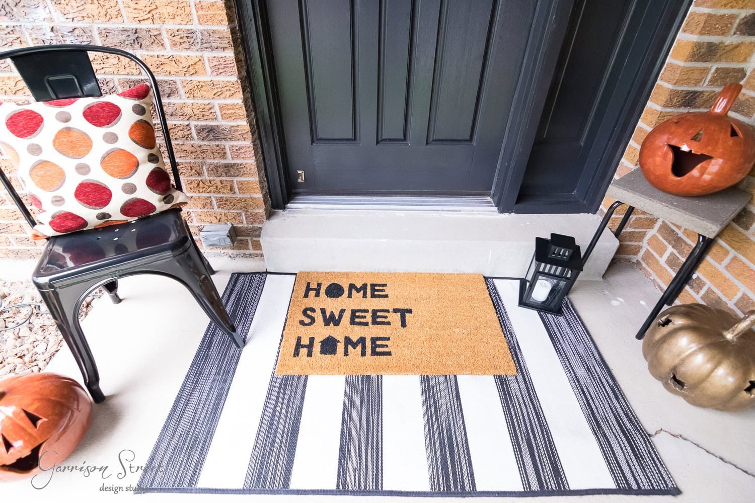 Front porch decorated for fall with a black door, a doormat reading "Home Sweet Home" on a striped rug, a chair with a polka dot cushion, carved pumpkins, and a lantern.