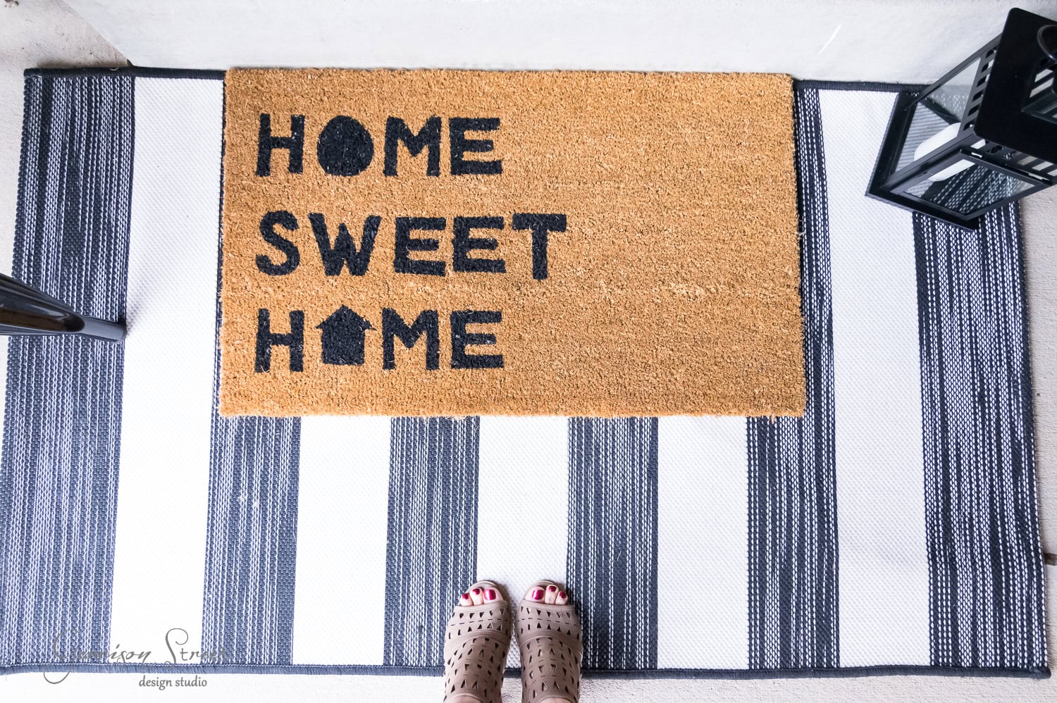 A doormat with the phrase "Home Sweet Home" is placed on a striped rug. Two feet wearing sandals are visible at the bottom edge of the image, and there's a black lantern on one side.