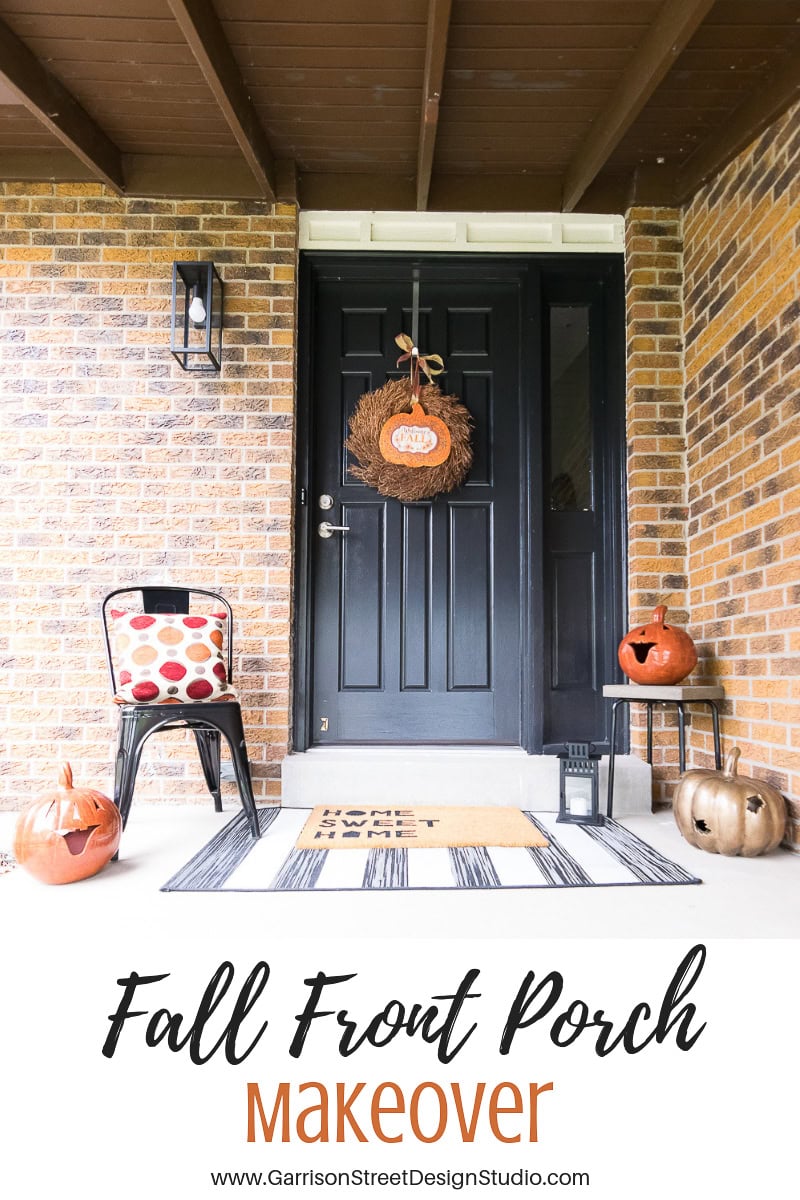 A black front door decorated for fall with a wreath and pumpkins on a porch. There's a black chair with a red and white polka dot cushion and a doormat. The porch has a brick wall background.