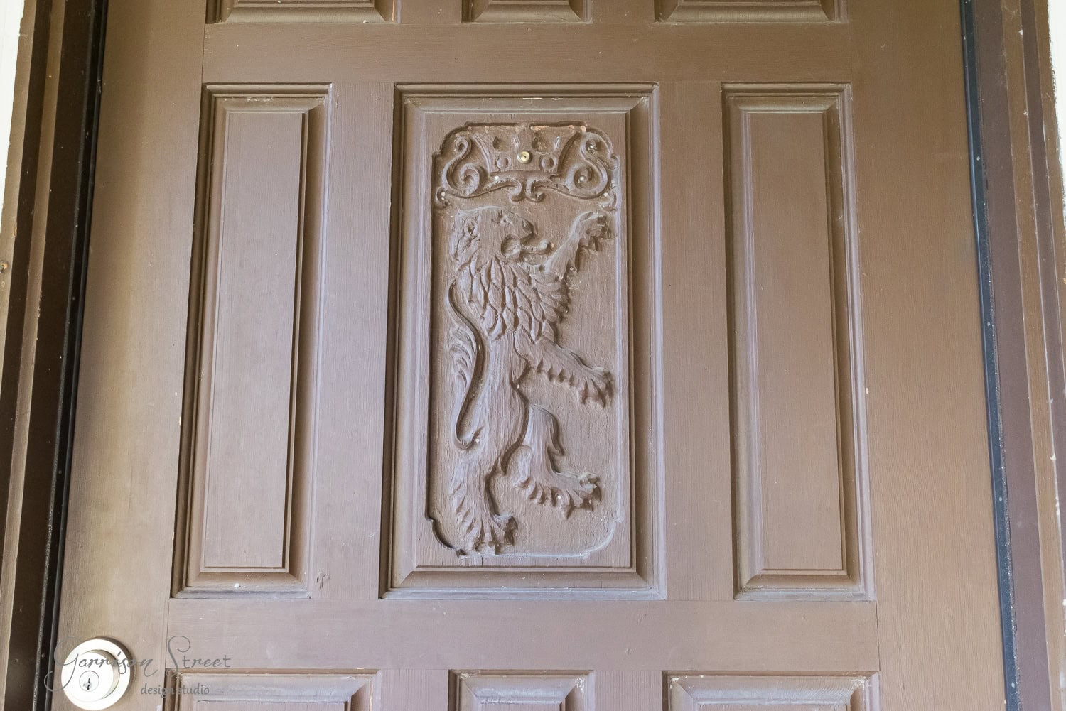 A brown wooden door with an intricate carving of a lion holding a staff and a crown above it. The carving is framed within a rectangular panel in the center of the door.