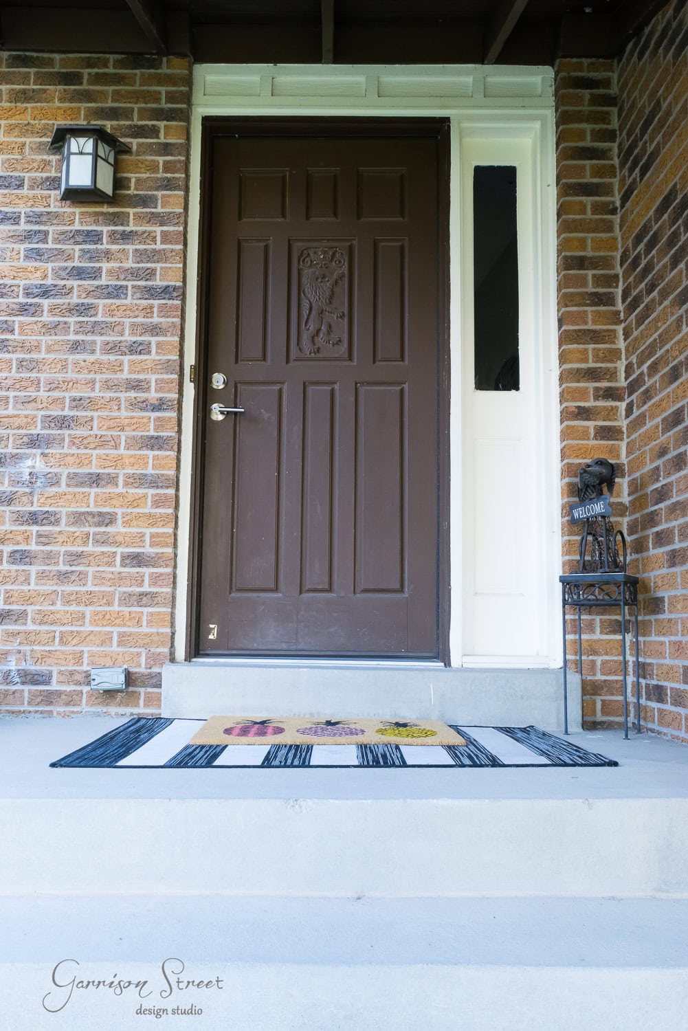 A brown front door on a brick house with a black porch light and white trim. A colorful welcome mat is on the step, and a small metal stand with a "Welcome" sign is on the right. The words "Garrison Street Design Studio" are in the lower left corner.
