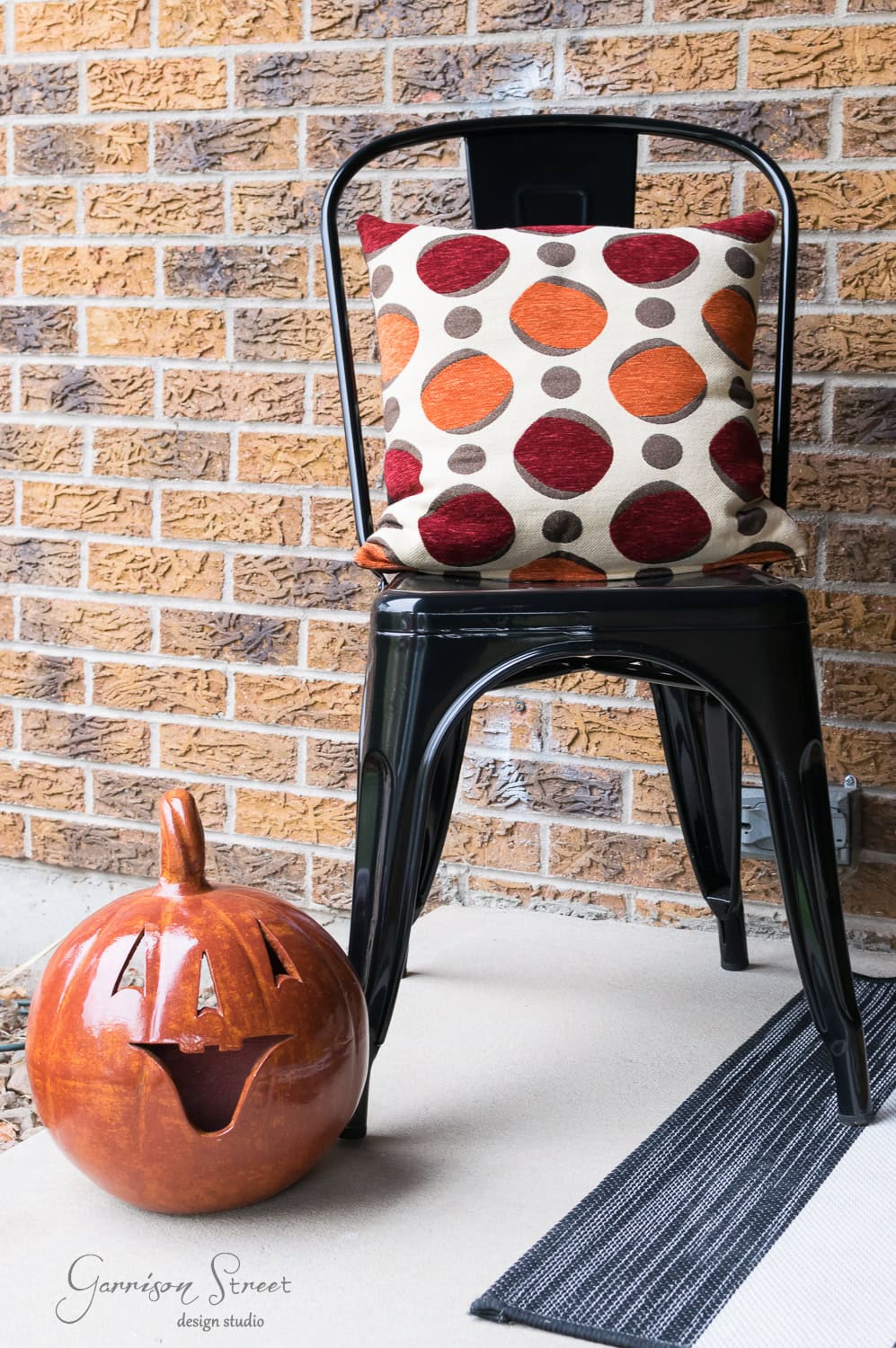 A black metal chair with a cushion featuring red and orange polka dots sits on a porch. Next to the chair is a smiling pumpkin decoration. The background is a brick wall and the floor has a black and white striped mat.