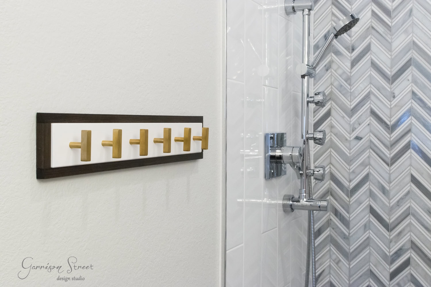 A sleek towel rack with gold hooks is wall-mounted beside the shower, showcasing a metallic showerhead and chevron-patterned tile. White tiles complement the space, while the Garrison Street design studio logo graces the corner. Ideal for any Ikea-inspired bathroom makeover.