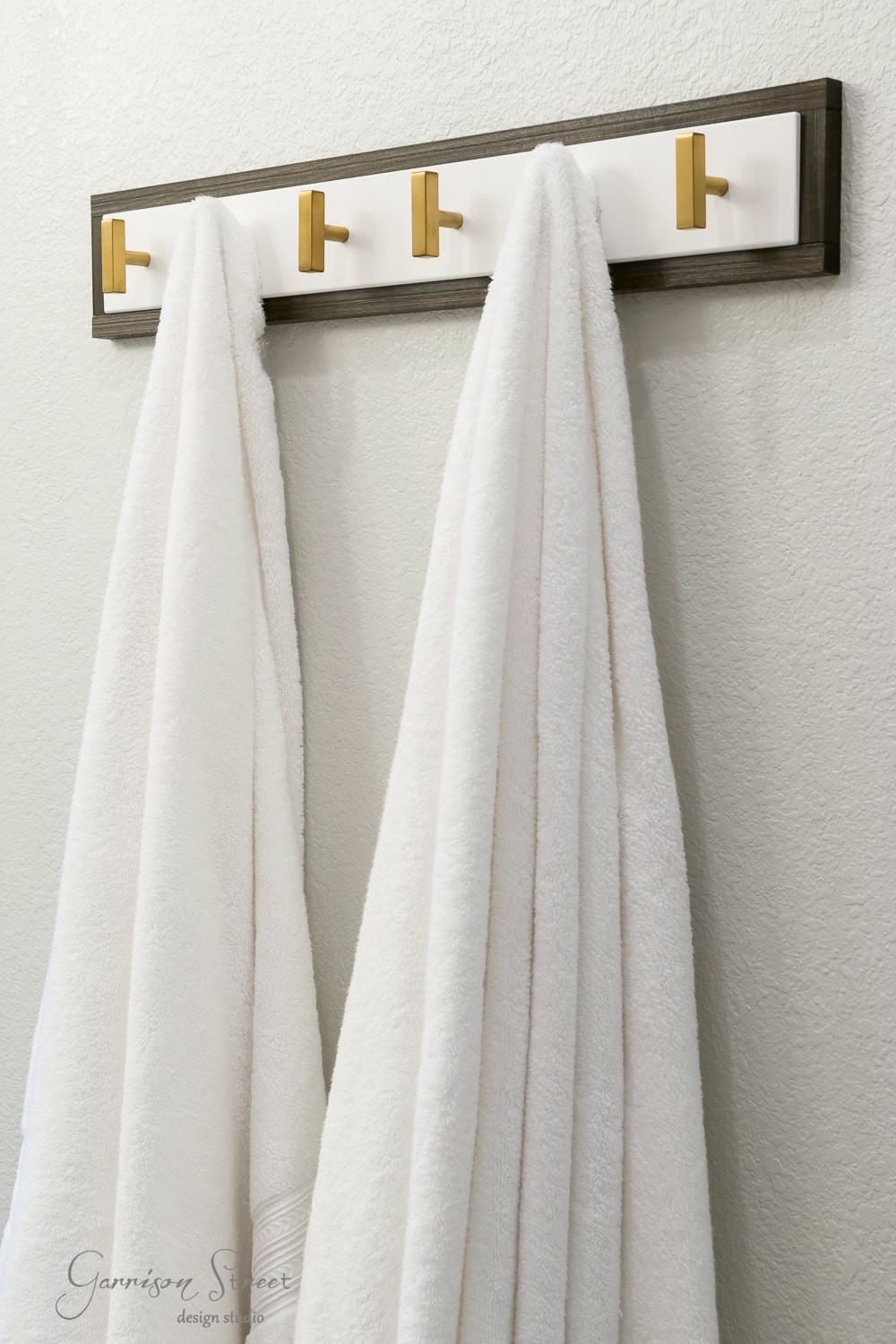 The Lurt towel rack, available at Ikea, features gold hooks that elegantly hold two white towels against a textured light-colored wall.