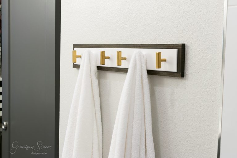 A modern Ikea towel rack featuring four gold hooks, each holding a white towel, is elegantly mounted on a light gray wall. The Lurt design includes a dark wood border and a white background. A gray door is partially visible to the left, complementing the sophisticated setup.