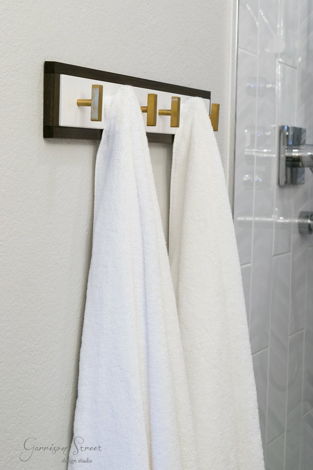 Two white towels hang on a sleek Ikea towel rack with gold hooks in a bathroom. The background showcases a neutral wall and a glimpse of beautifully tiled shower walls.