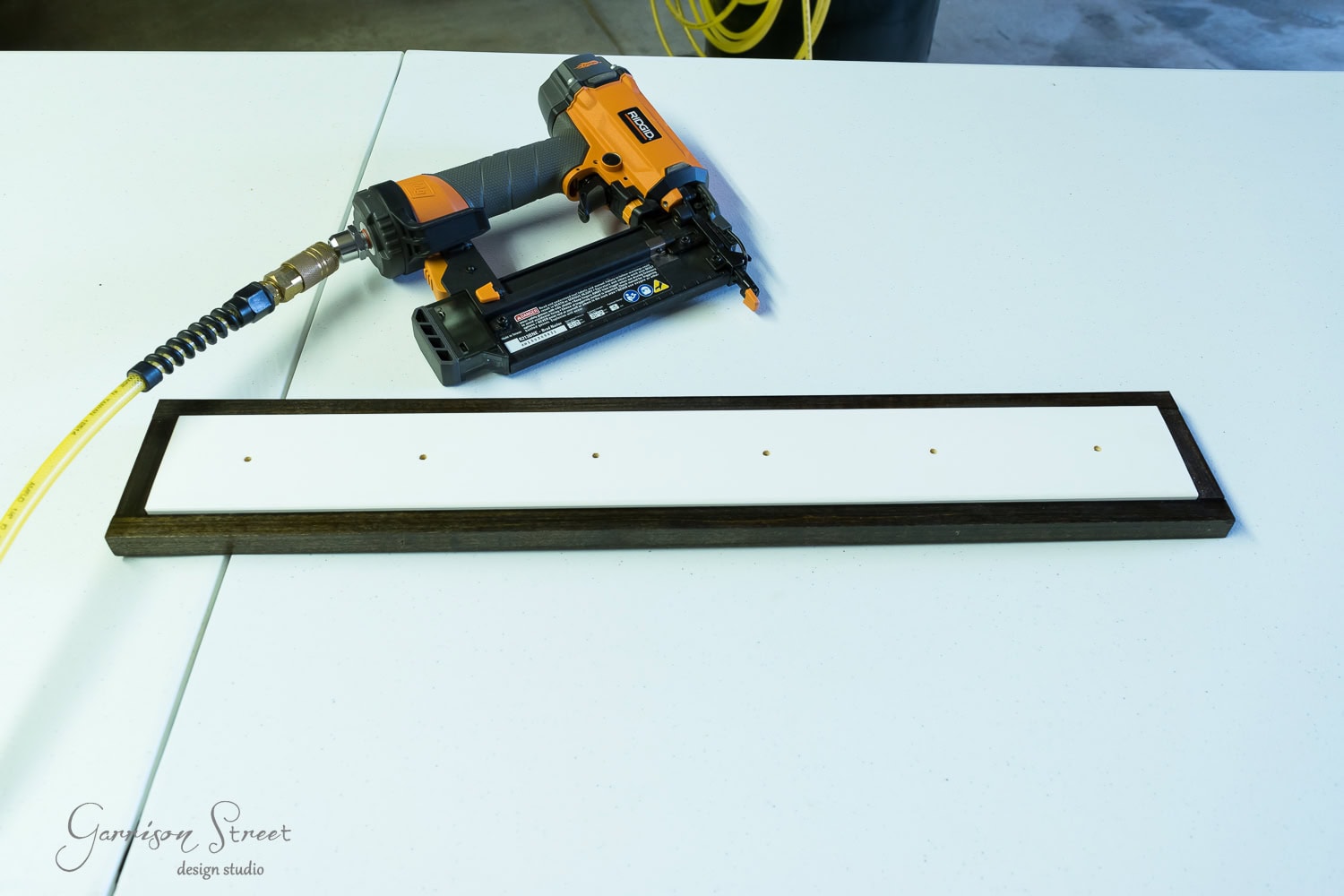 A nail gun is placed next to a wooden board on a white table, possibly for assembling an Ikea towel rack. The board, framed in dark brown with five small holes, suggests a woodworking or DIY project in progress. The nail gun is connected to a yellow air hose.