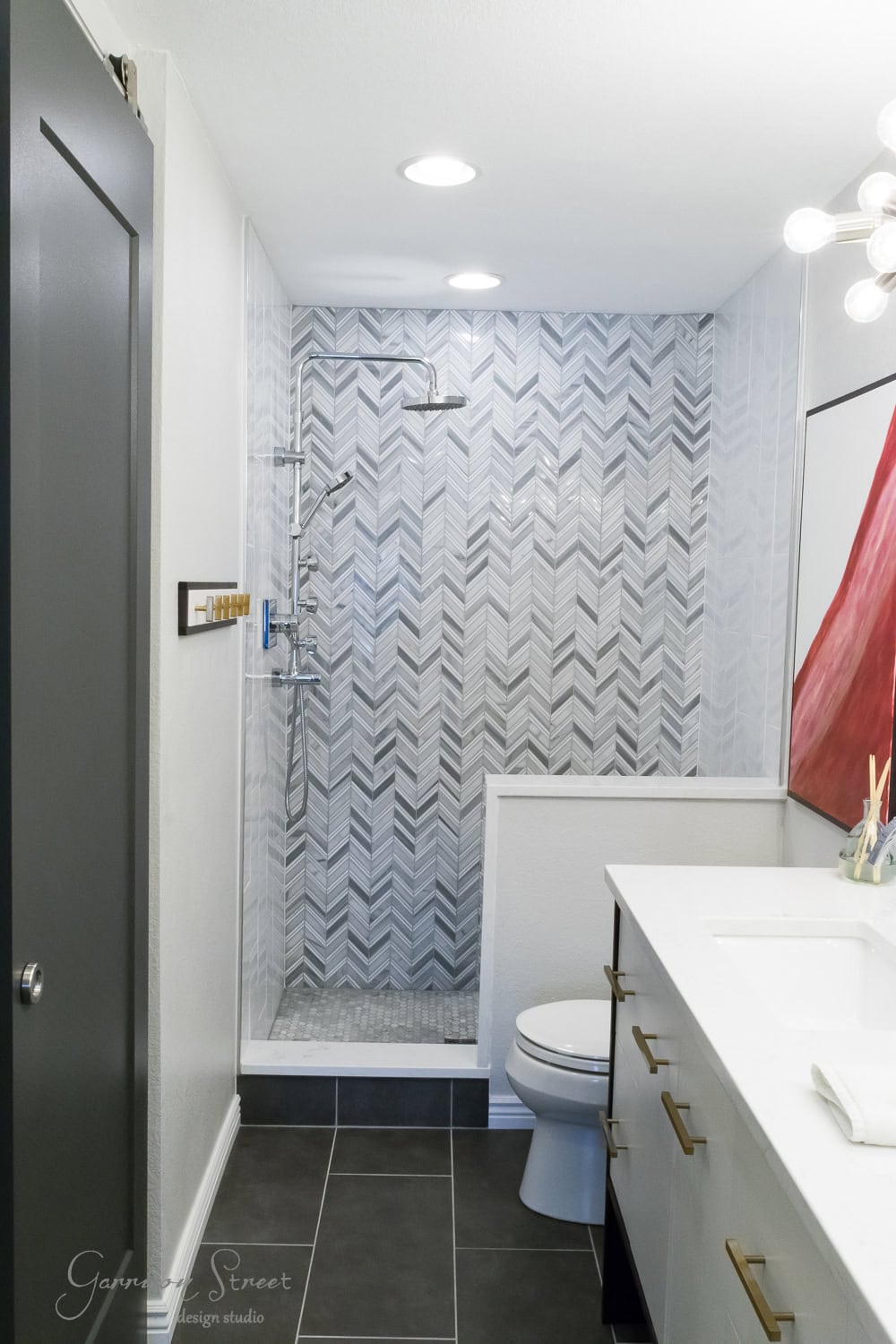 Modern bathroom with a herringbone-patterned tiled shower, glass door, white sink counter, and a gray floor. There's a rectangular mirror with light fixtures above and abstract art on the wall.