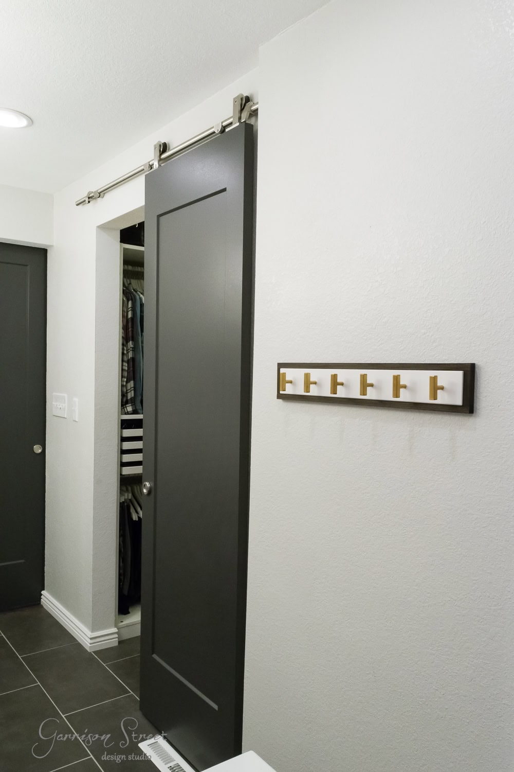 Minimalist hallway with a sliding grey door revealing a closet. The closet has neatly arranged clothes. On the wall, there's a row of gold hooks. The floor is covered with dark tiles, and the room appears brightly lit.