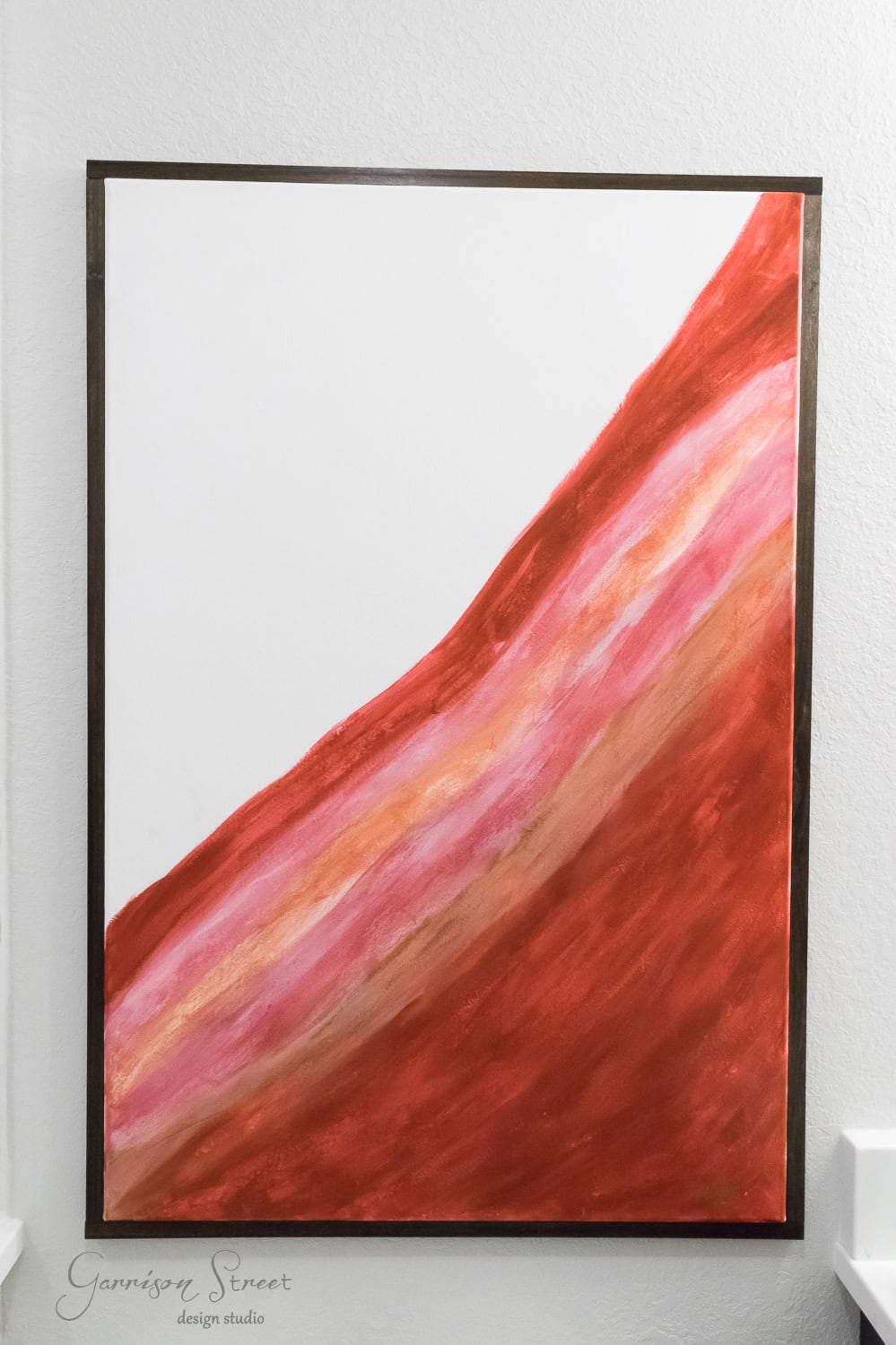 Abstract painting with a diagonal sweep of earthy tones, including red, pink, and orange, resembling layers of sediment. The border is thin and dark, set against a white background. The artwork appears to be signed in the lower left corner.