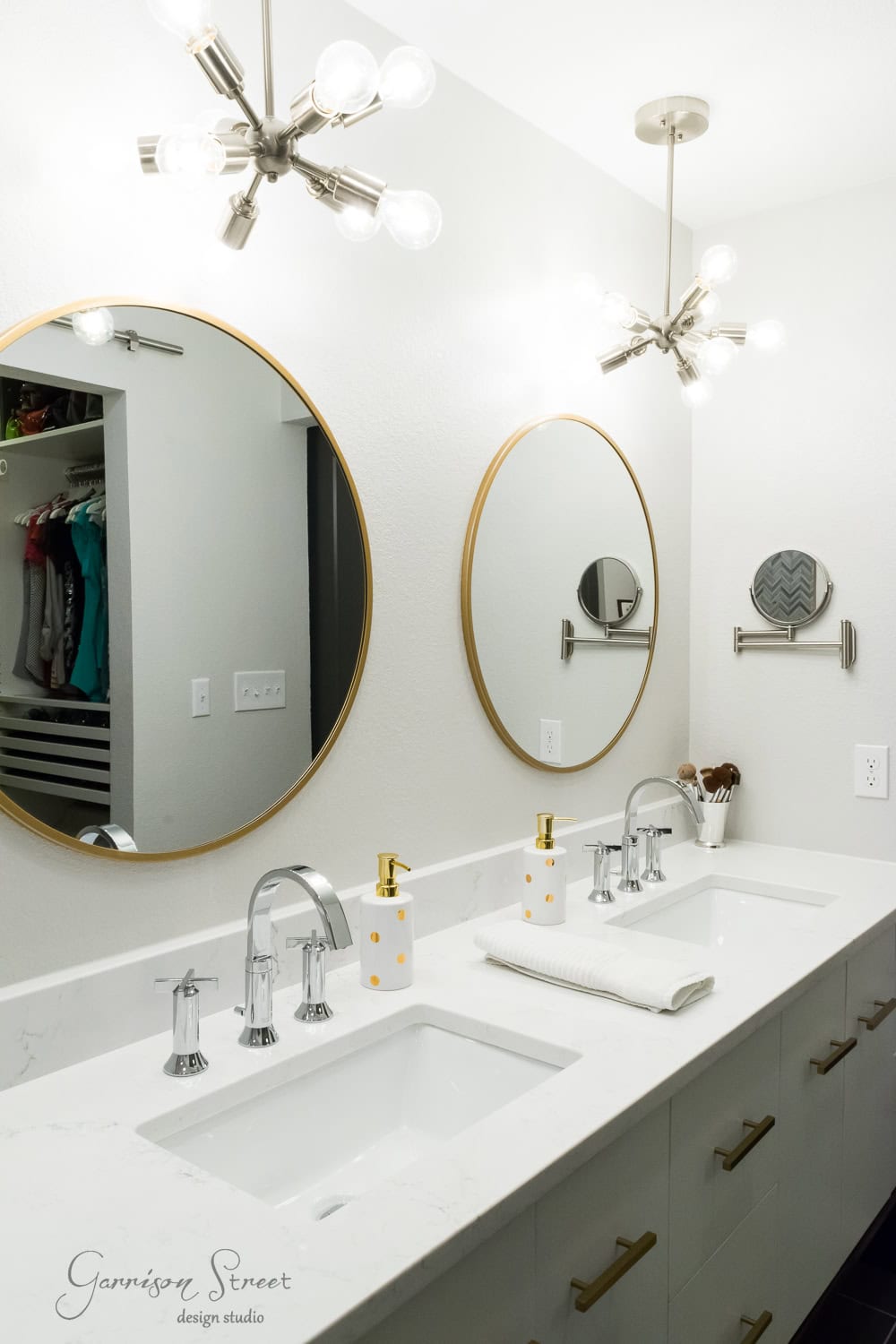 A modern bathroom vanity with two round mirrors, brass fixtures, and a marble countertop. Two sinks are equipped with sleek faucets, accompanied by soap dispensers and neatly folded towels. Ceiling lights provide bright illumination.