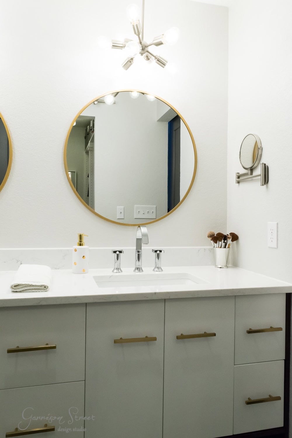 Modern bathroom with a white vanity, featuring brass handles and a quartz countertop. A round mirror with a gold frame hangs above the sink, and a smaller magnifying mirror is mounted on the wall. Overhead lighting has a sleek, contemporary design.