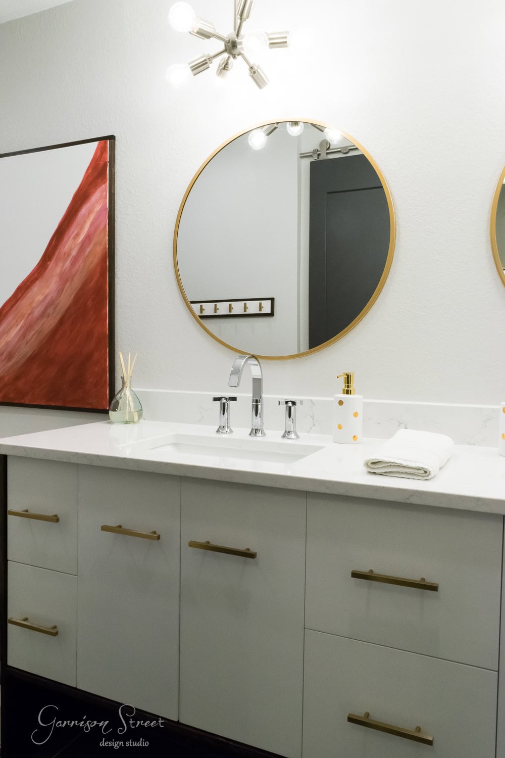 Modern bathroom with a white vanity, gold handles, and two round mirrors. A contemporary light fixture hangs above. A large abstract painting with red hues is on the left wall. A white hand towel and a small container sit on the counter.