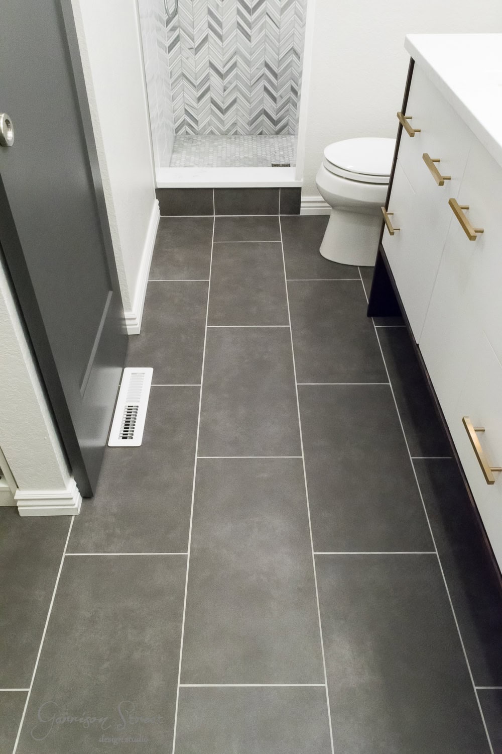 Modern bathroom with large dark gray tiles on the floor. A white toilet sits beside a white vanity with gold handles. The shower features a chevron-patterned tile wall. A gray door is partially visible on the left.
