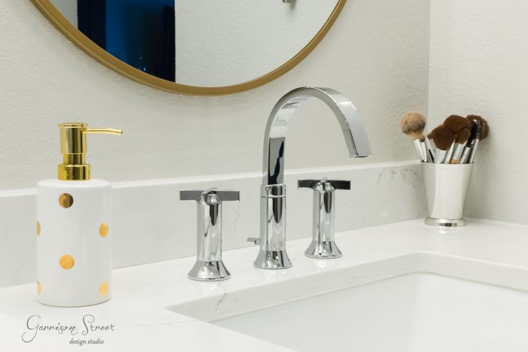 A modern bathroom sink with a sleek chrome faucet. A white and gold polka dot soap dispenser and a cup holding makeup brushes sit on the white countertop. A round mirror with a gold rim is partially visible.