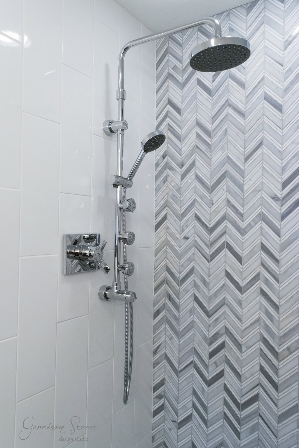 Modern shower with dual heads and adjustable handheld, mounted on a white and gray herringbone-patterned wall. The fixtures are sleek and metallic, complementing the tiled design.