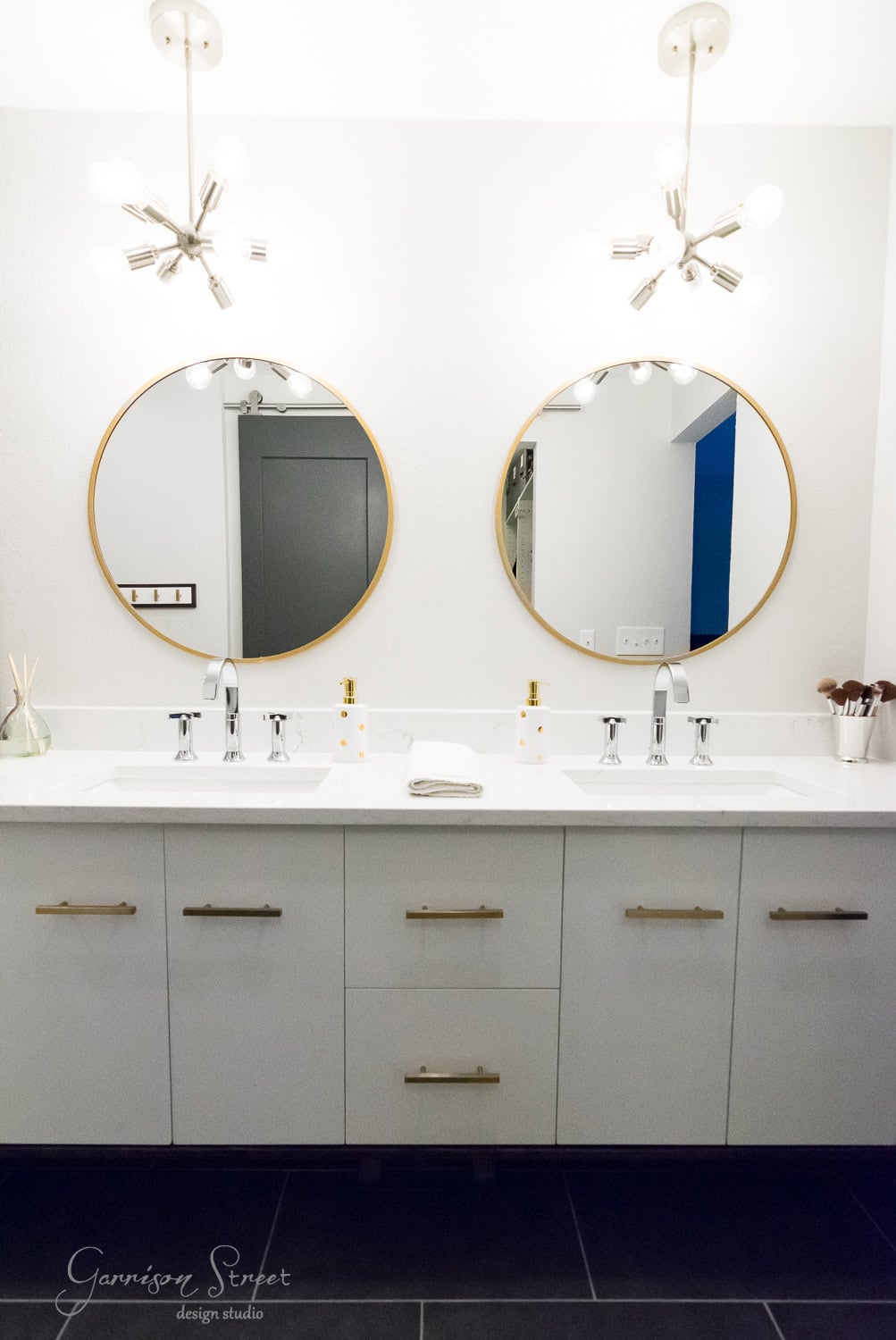 A modern bathroom with a double vanity featuring round mirrors, white sinks, and gold accents. Overhead are two stylish light fixtures. The cabinetry is white with gold handles, and the floor is dark.