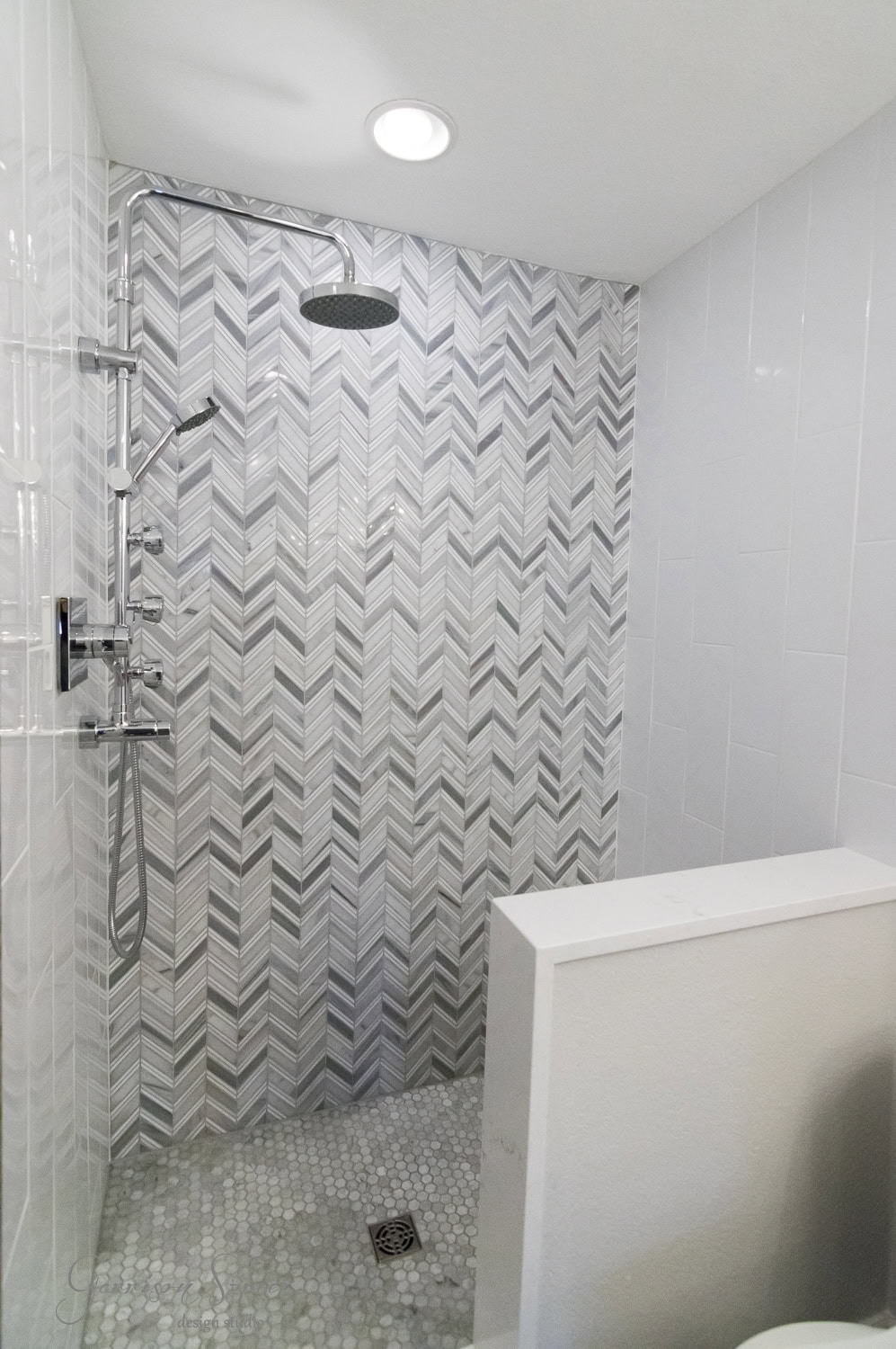 A modern shower with a herringbone tile pattern in shades of gray and white. The floor features small circular tiles. A large overhead rain showerhead is mounted on the wall, with additional controls and a handheld shower wand.