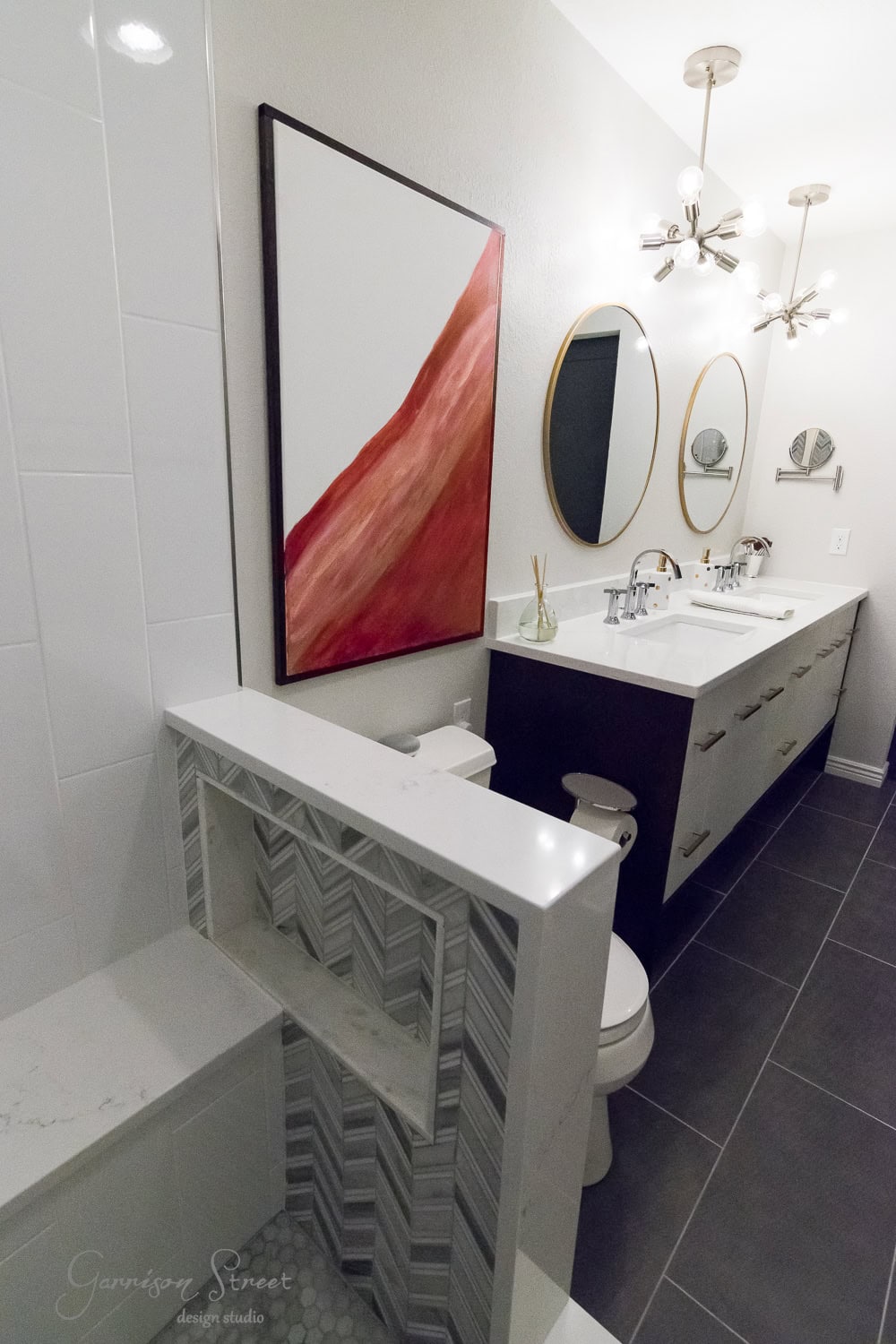 Modern bathroom with a large red abstract painting, two round mirrors above a double sink vanity, geometric pendant lights, and a mix of dark and light tile surfaces.