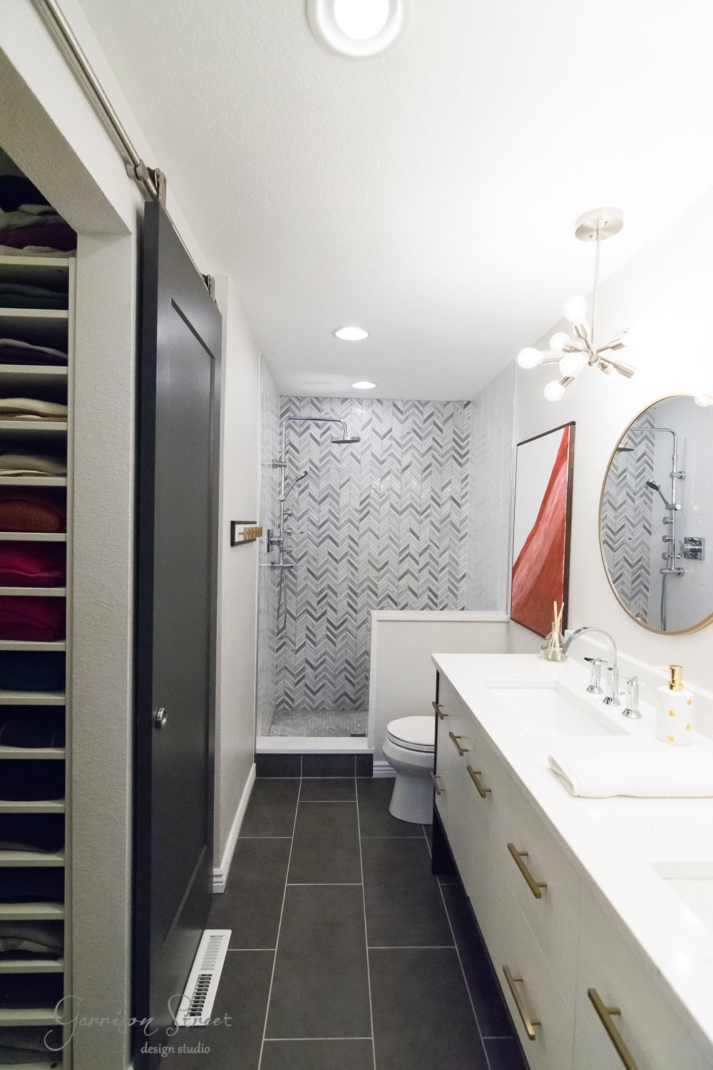 A modern bathroom with a sliding door reveals a shower area lined with chevron-patterned tiles. The space features a double sink vanity with a round mirror and contemporary light fixture, alongside gray floor tiles and a small shelf with folded towels.