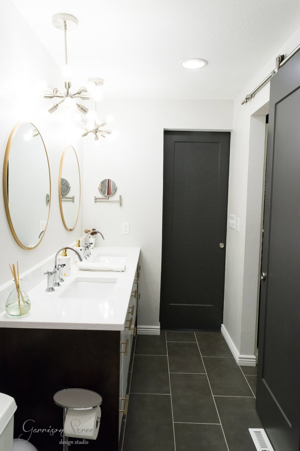 Modern bathroom with dark floor tiles, a double vanity with two round mirrors, and a sliding barn door. The space features a white countertop, chrome faucets, and decorative lighting. A small stool is in the corner.