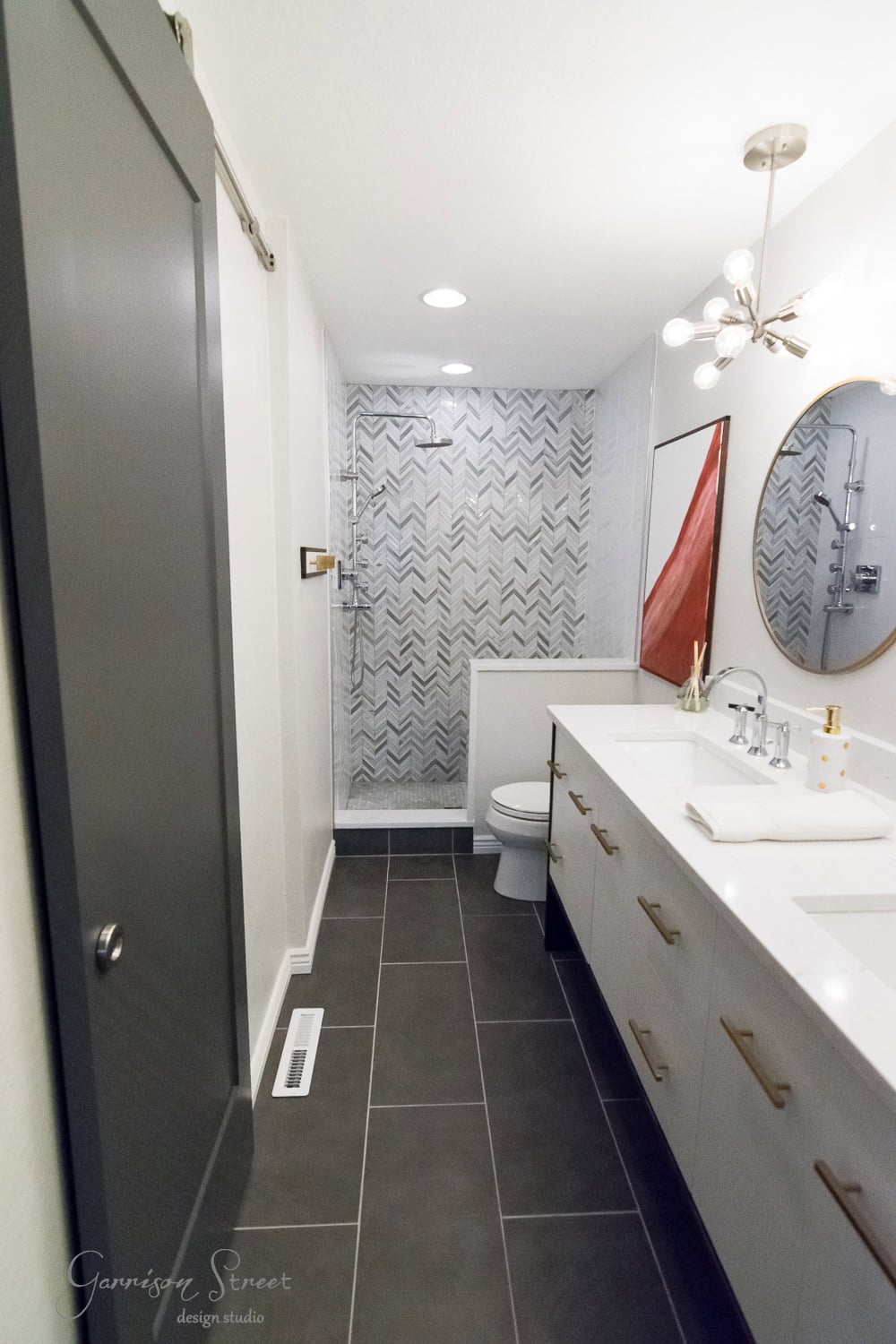 Modern bathroom with large tiles and a walk-in shower featuring herringbone-patterned tiles. A white vanity with dual sinks and gold accents is opposite a sliding barn door. A round mirror and a ceiling light add elegance.