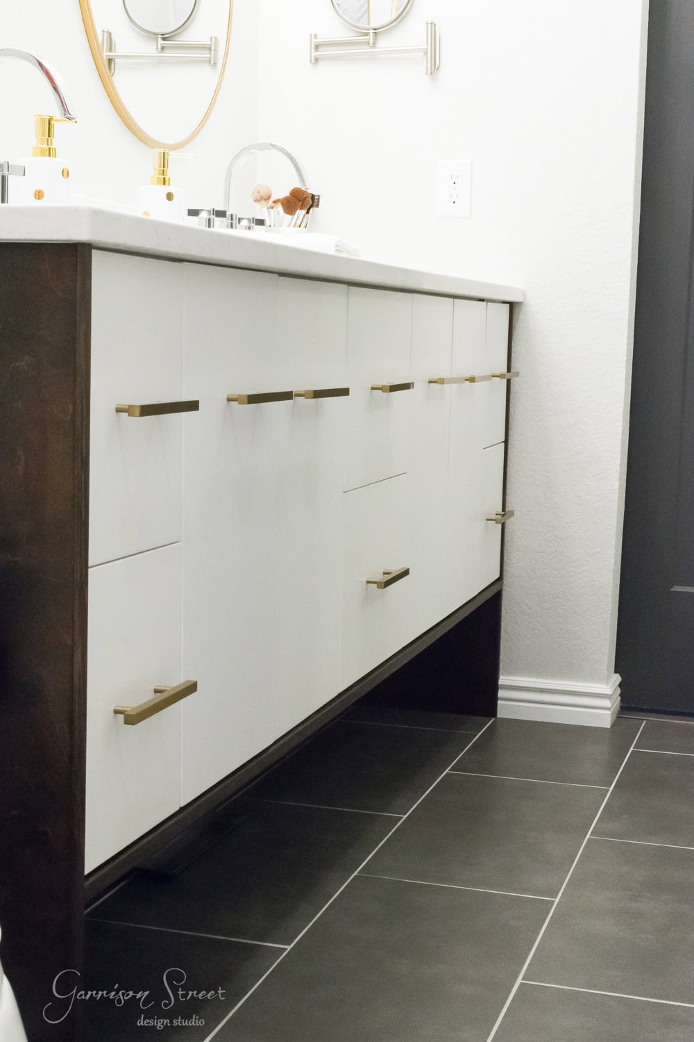 Minimalist bathroom with sleek white cabinetry, gold handles, and dark wood accents. Gray tile floor and circular mirrors reflect contemporary design. Simple decor includes a decorative round tray and glass dispenser on the counter.