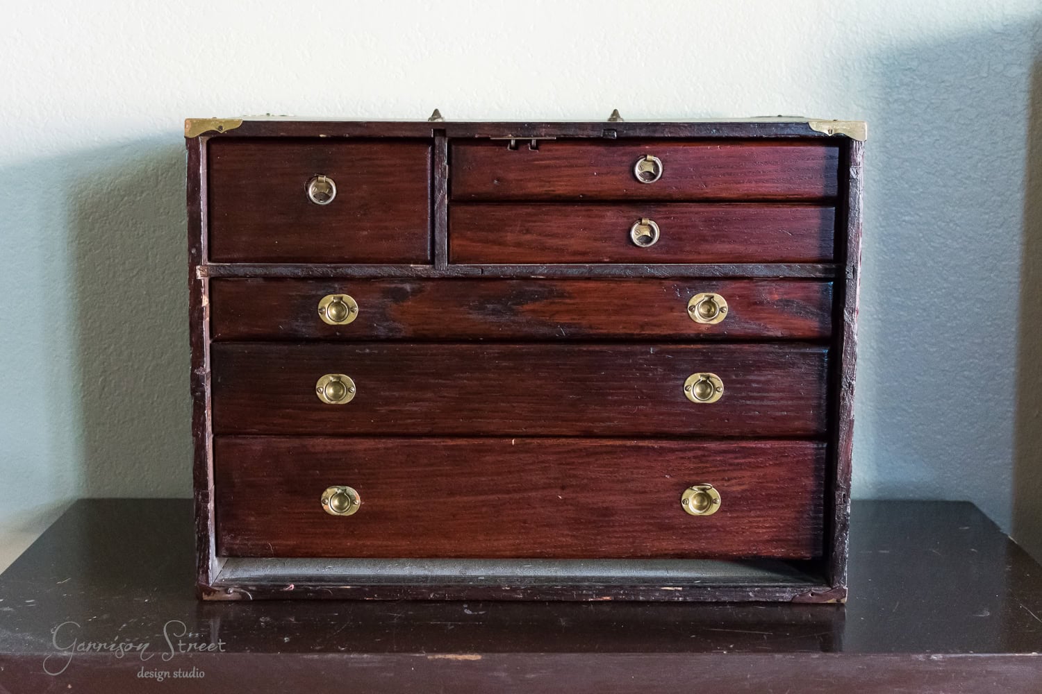 A vintage wooden tool chest with multiple drawers featuring brass handles. The chest boasts a dark, polished finish and rests elegantly against a light-colored wall on a flat surface.