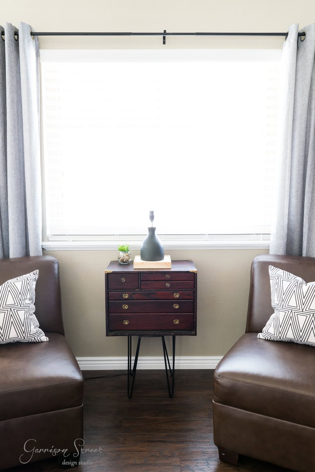 A cozy living room corner features two brown leather chairs, each with a geometric-patterned pillow. Nestled between them is a small wooden side table on metal legs, reminiscent of a vintage tool chest, holding a black vase and small plant. Gray curtains frame the sunlit window behind.