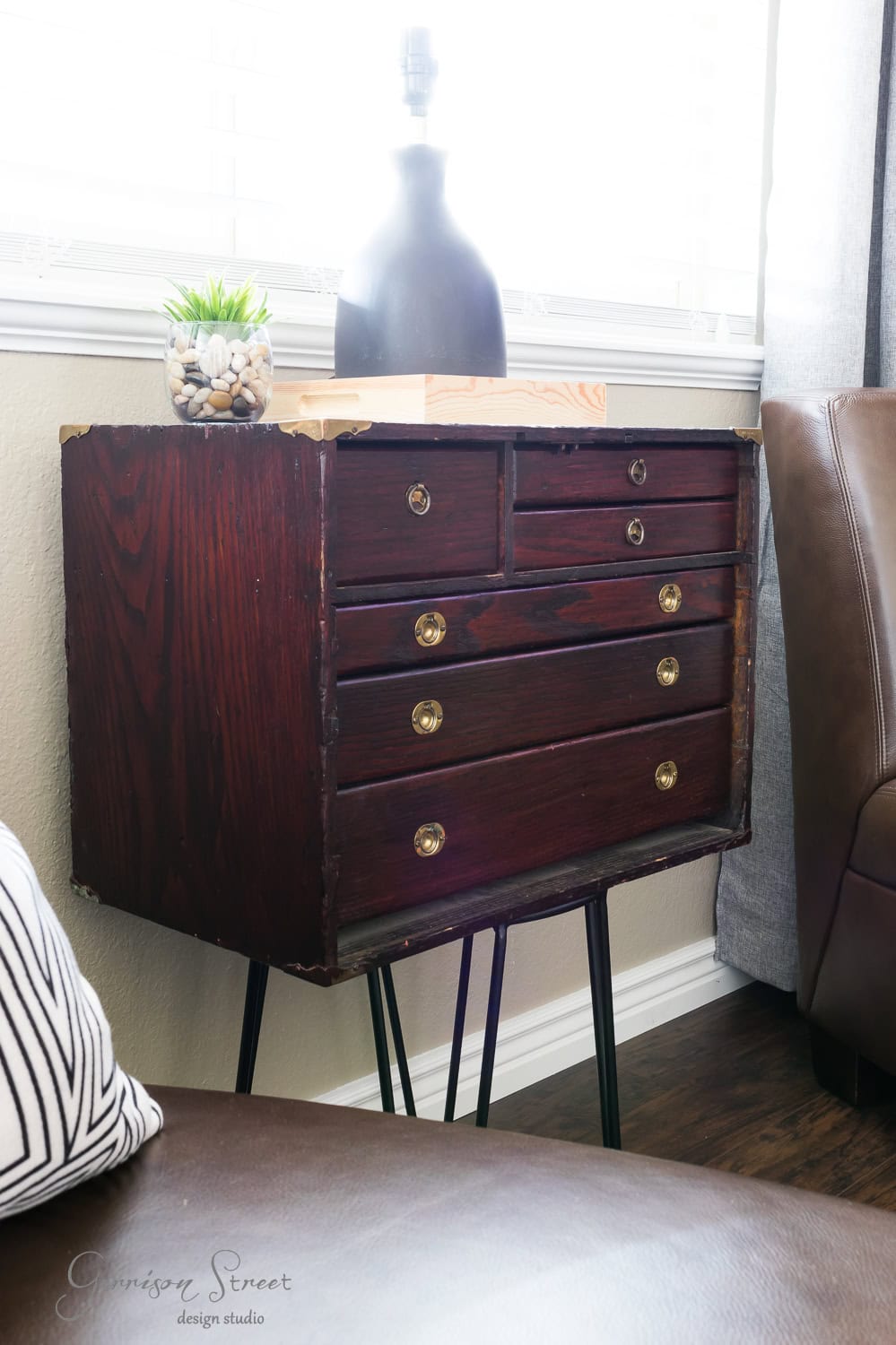 A vintage wooden cabinet with six drawers and round brass handles stands on black hairpin legs, reminiscent of an old-world tool chest. It's beside a brown leather chair, and a black lamp adorned with pebbles is on top. Soft light filters through venetian blinds in the background.