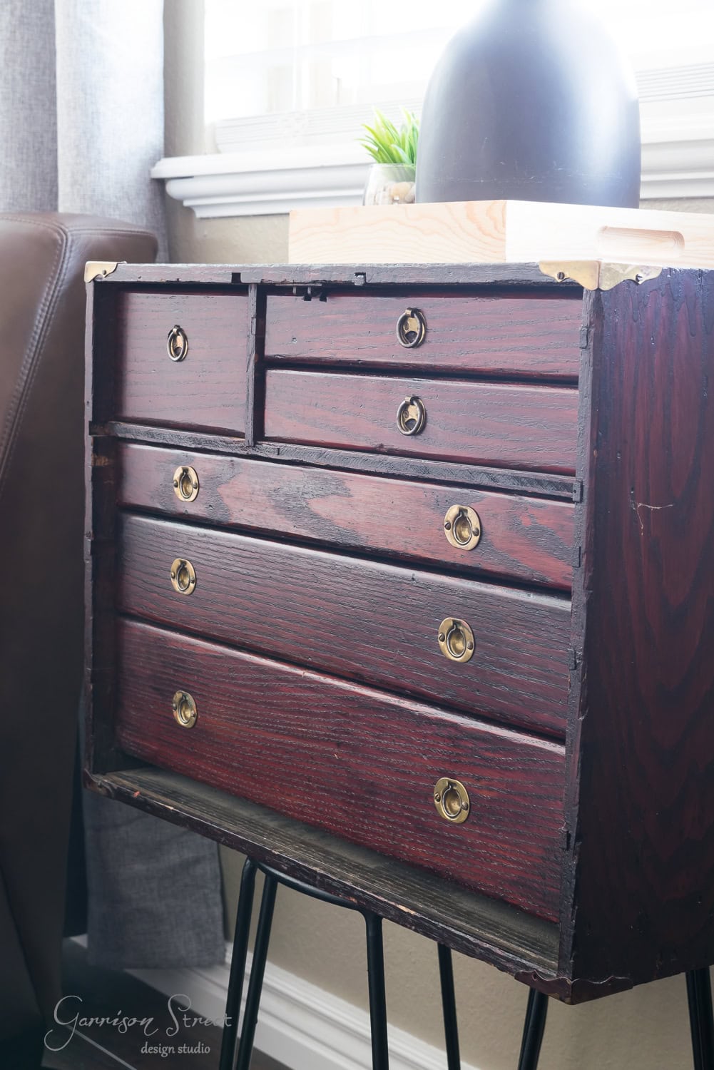 A vintage wooden dresser with multiple drawers, reminiscent of an old tool chest, features brass handles. It stands on metal legs in front of a window, adorned with a dark vase and small plant on top.
