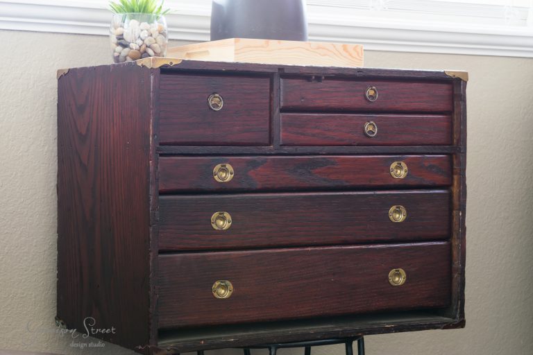 A vintage wooden dresser with six drawers and brass knobs stands against a wall. A plant and a black vase sit on top, placed in front of a window. The dresser has a worn, rustic appearance.