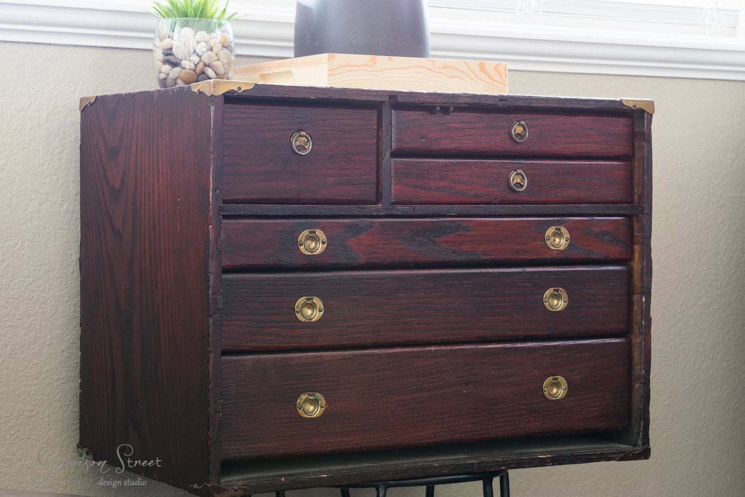 A vintage wooden dresser with six drawers and brass knobs stands against a wall. A plant and a black vase sit on top, placed in front of a window. The dresser has a worn, rustic appearance.